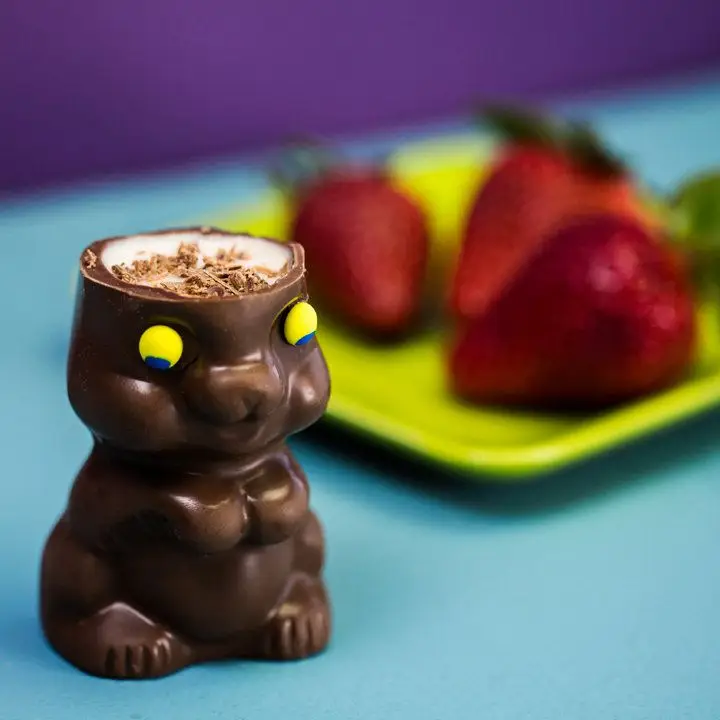 In the foreground, a chocolate Easter bunny with yellow eyes and no top of its head is filled with white liquid and dusted with chocolate. In the background, out of focus, is a yellow plate with three fresh strawberries on top.