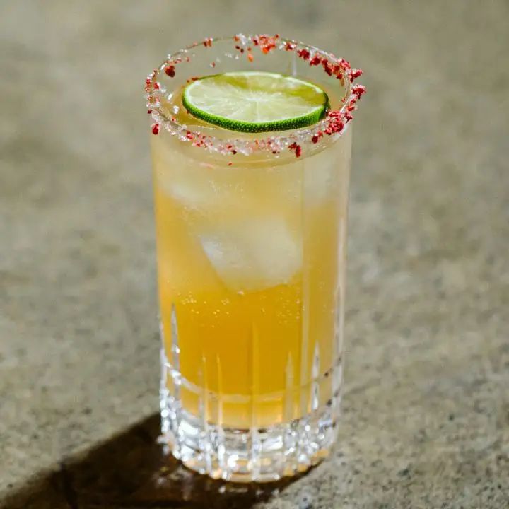 On a gray surface, a faceted high ball glass is filled with an orange-hued soda and a few ice cubes. Its rim is dusted with bright red flakes, and it’s garnished with a single lime wheel.
