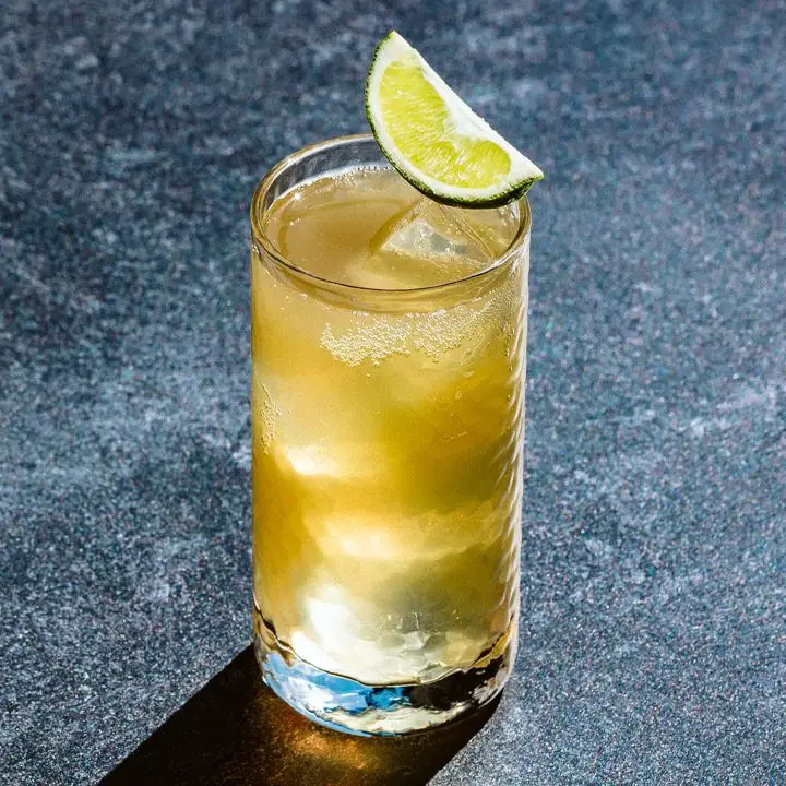 A textured highball glass containing a golden effervescent cocktail garnished with a lime wedge