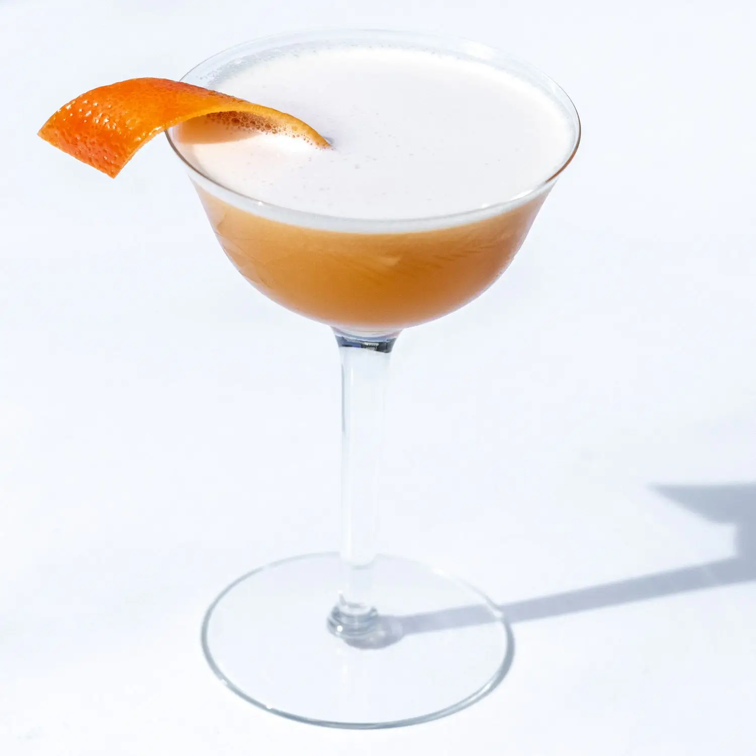 Brown Derby cocktail in high-stemmed coupe glass, with a head of foam and orange twist, on a light colored background