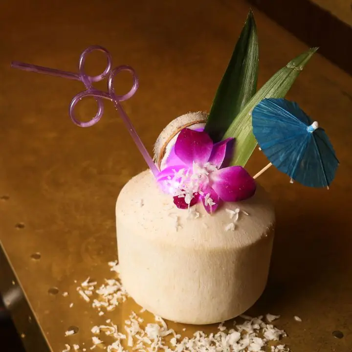 A peeled coconut shell rests on a copper bar top. It’s filled with a bright pink flower, a curly pink plastic straw, a blue paper umbrella, two coconut fronds and shavings of coconut.