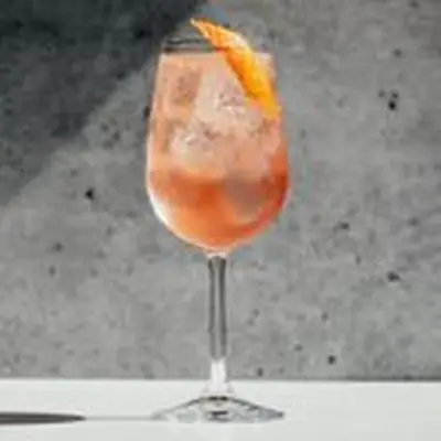 A tall wine glass stands on a white surface against a concrete backdrop. The glass is filled with a rose-colored sparkling drink, ice cubes and an orange peel.