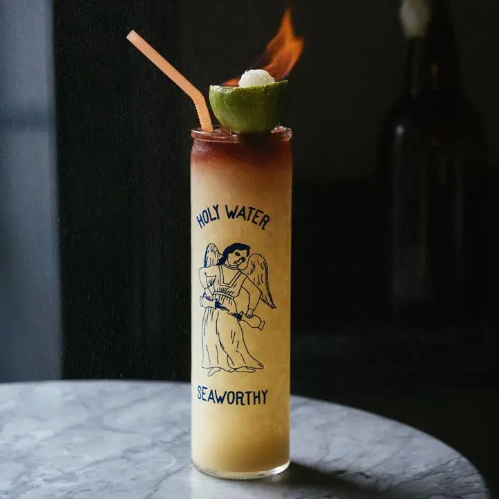 A tall highball glass sits on a marble table. It reads Holy Water Seaworthy, with a drawing of an angelic figure. It’s filled with crushed ice and light brown drink, with a dark float on top. Atop that is a lime half with a burning sugar cube, as well as an orange bendy straw.