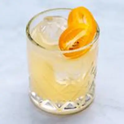 A tiny rocks glass with diamond etchings sits on a marble surface. The drink within is yellow and garnished with a halved kumquat.