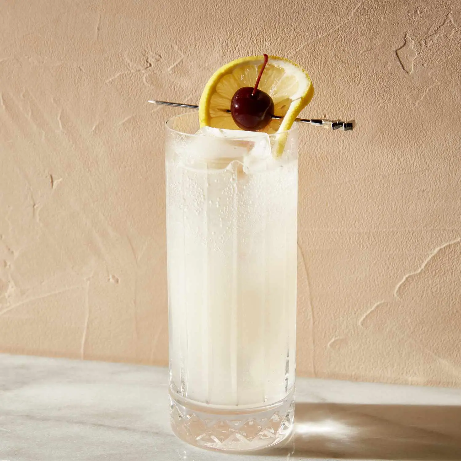 Tom Collins cocktail in tall, narrow Collins glass, with lemon wheel and cherry garnish on top 