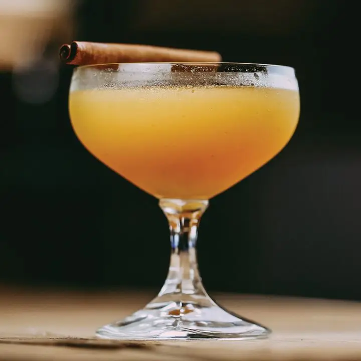 orange-hued comrade cocktail in a short-stemmed coupe glass, garnished with a cinnamon stick balanced on the rim