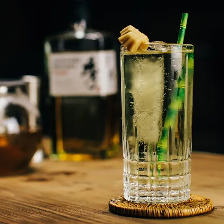 In the foreground, a Collins glass with a faceted bottom rests on a wicker coaster. It’s filled with a green highball, a spear of ice, a fake bamboo straw and a maple leaf candy garnish. In the background out of focus is a bottle of Japanese whisky.