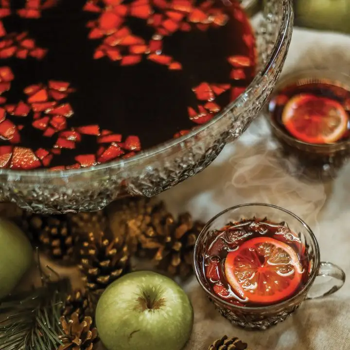 A large glass bowl filled with dark red punch rests on a white tablecloth, surrounded by gold pinecones, green apples and glass mugs of punch.