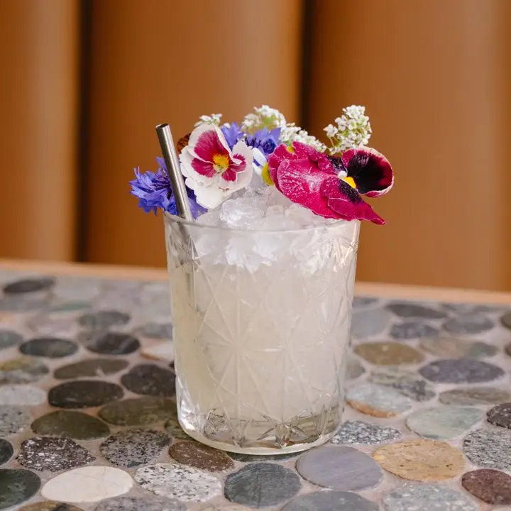 Rebirth cocktail in a textured rocks glass, served over crushed ice and garnished with purple, pink and white flowers and a metal straw