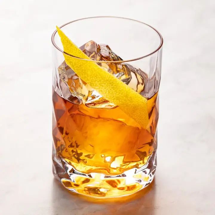 A tall, narrow rocks glass holds some ice cubes and an orange-hued drink. A long, angular piece of lemon peel garnishes the drink, and the whole thing is set against white marble.