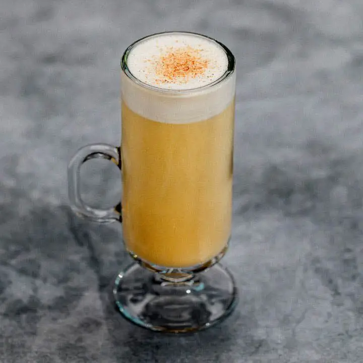 A tall, thin Irish Coffee mug rests on a dark gray marbled surface. The glass mug holds a pale gold drink with a thick layer of white foam, dusted with nutmeg, at the top.