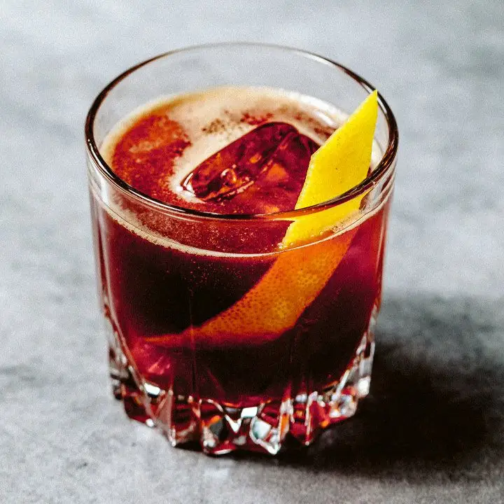 A rocks glass rests on a gray marble surface. The glass is filled with a dark red opaque drink over a large ice cube, and is garnished with a thin slice of lemon peel.