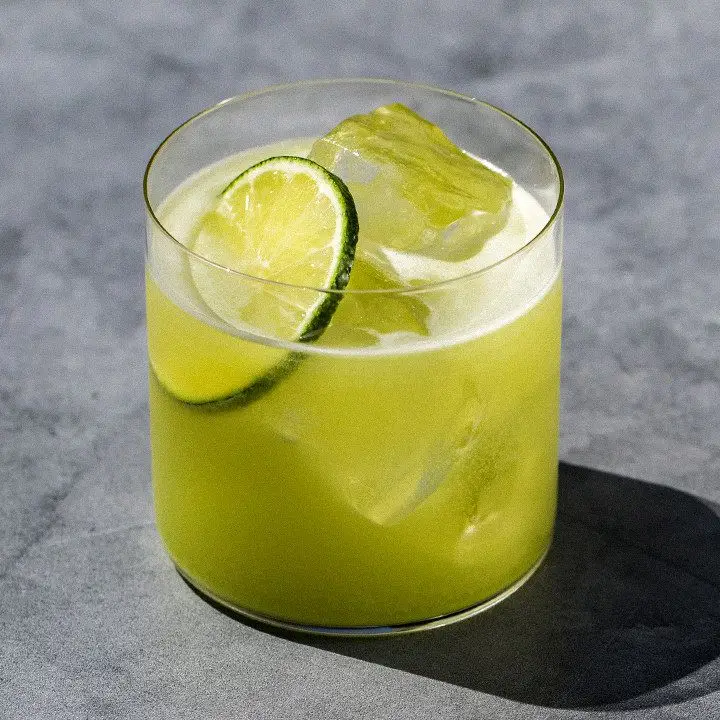 A thin-walled rocks glass sits on a gray concrete surface. The glass holds a vivid green cocktail over ice, garnished with a lime wheel.