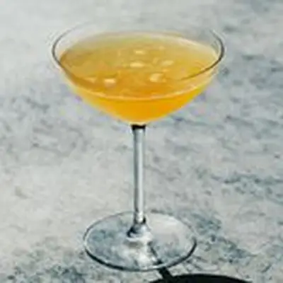 A narrow-stemmed cocktail glass with a wide brim holds a pale orange cocktail. A few bubbles on the surface reveal how recently it was poured, and the orange of the drink is set off against the gray marble backdrop.