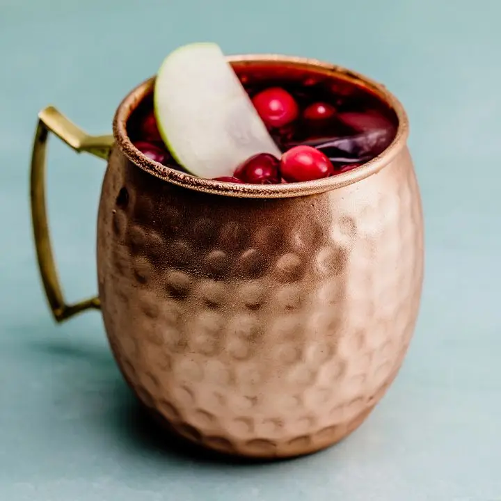 A copper mug with a handle and hammered finish sits on a blank, blue backdrop. The mug is filled with ice, and garnished with fresh ripe cranberries and a slice of green apple.