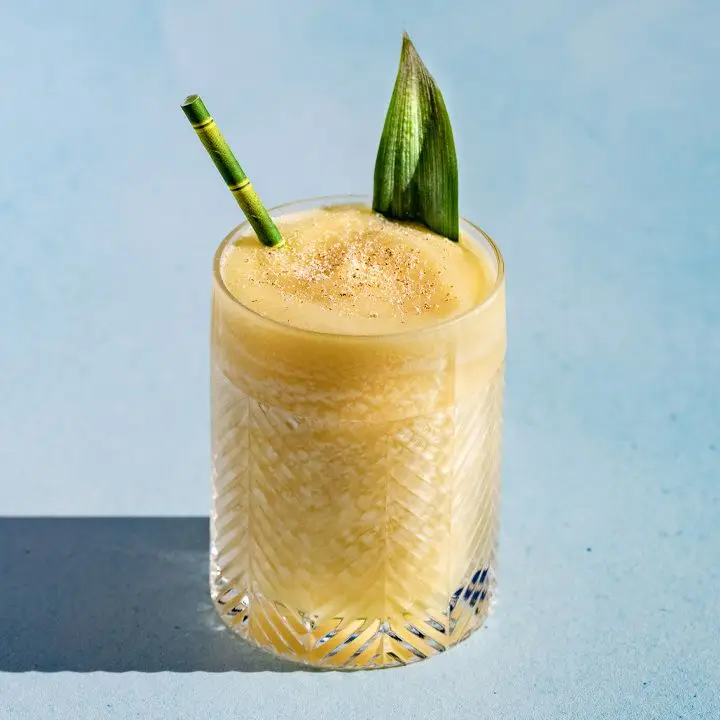 A double rocks glass rests on a light blue surface. It’s filled with a creamy blended yellow drink and garnished with a pineapple frond and a bamboo-colored straw.
