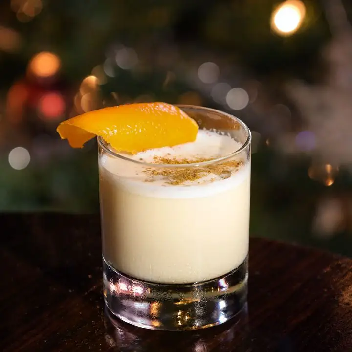 A creamy Eggnog drink fills a rocks glass with a heavy clear base. The drink is garnished with nutmeg and an orange peel, and the background is hazy.