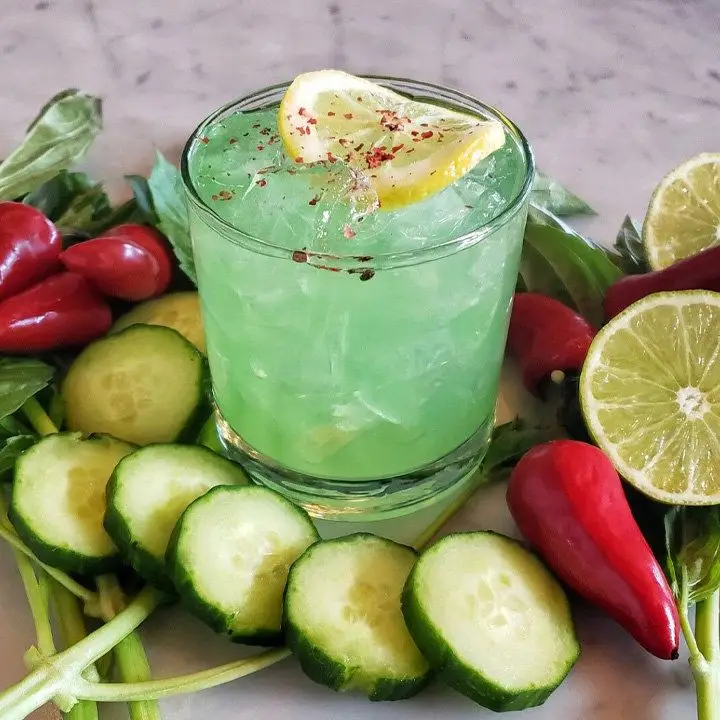 bright green Elder Greene Blues cocktail in a rocks glass, with lemon wheel garnish, surrounded by sliced cucumbers and red peppers