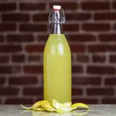 A swing-top bottle sits on a marble countertop in front of a brick wall. The bottle is filled with a bright yellow liquid, and a number of lemon peels are clustered in front of it. 