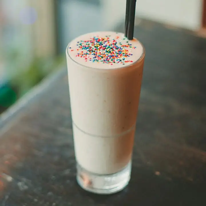 A tall, thin glass holds a frothy pale drink, its surface dotted with brightly colored round sprinkles. The drink has two black bar straws and rests on a wooden table. 