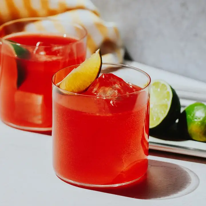 Two thin-walled, thin-based rocks glasses rest on a white surface. They’re filled with a bright red drink, ice and lime wedges. Cut limes rest on a plate behind them, along with a yellow and white-striped towel.