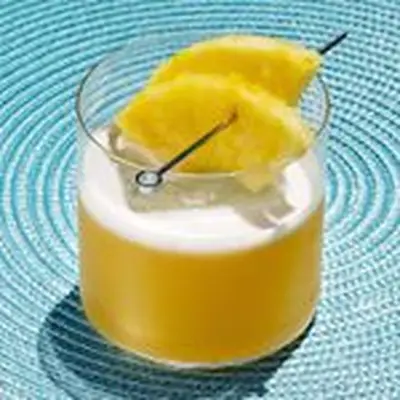 A thin based rocks glass rests on a sky blue ringed mat. The glass holds a large ice cube, a golden drink and two pineapple wedges on a silver skewer.