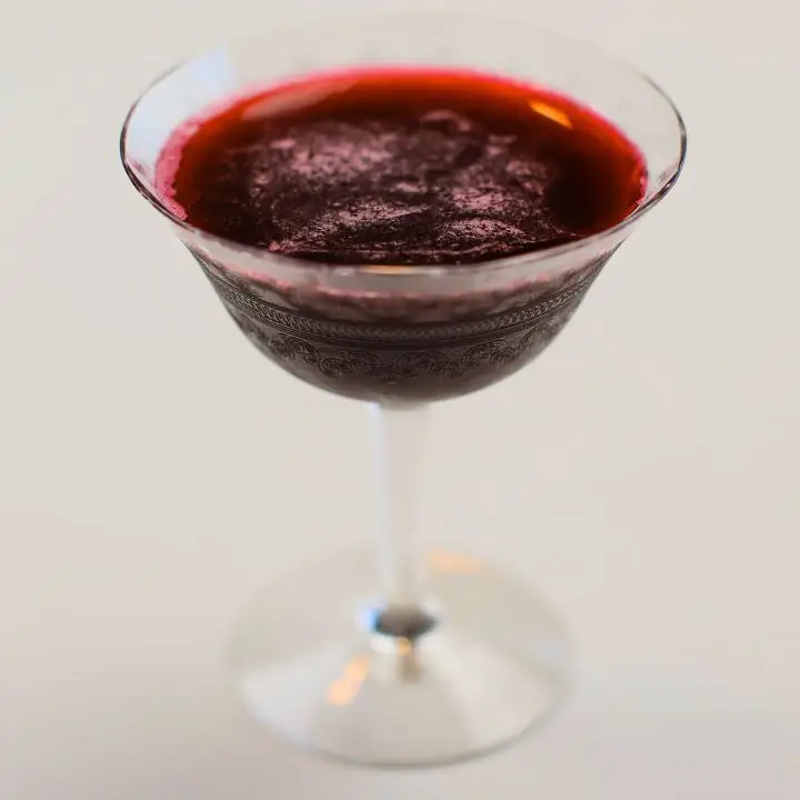 A beautifully etched cocktail glass holds a blood-red drink on a white backdrop.