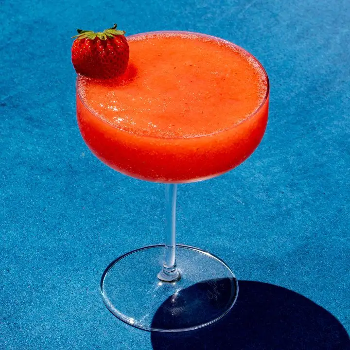 A vibrant orange-pink-colored, blended Strawberry Margarita cocktail in a stemmed cocktail glass with a strawberry on the rim for garnish.