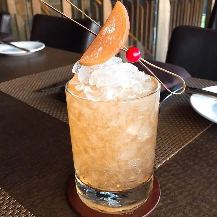 A peach-colored cocktail served in a rocks glass over crushed ice and garnished with a pickled peach slice on a retro wooden pick