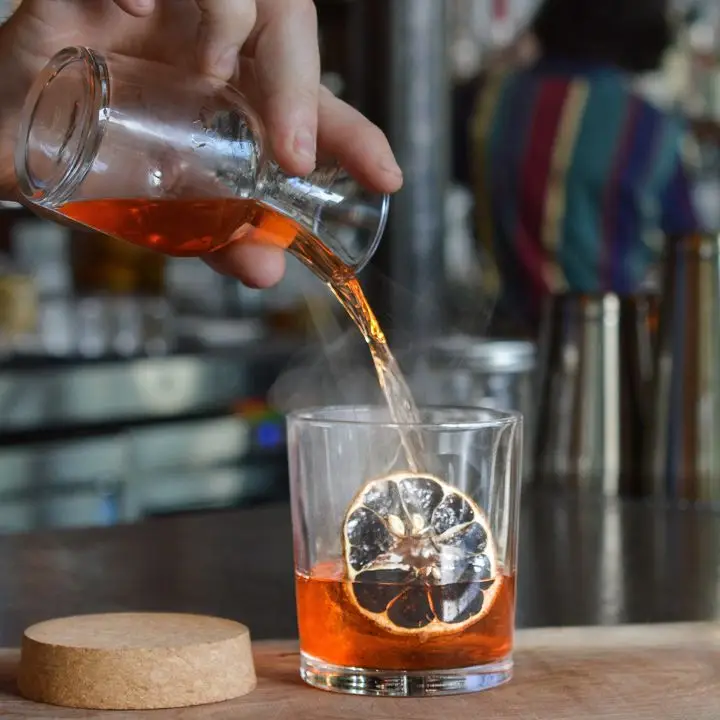 A bartender pouring a ruby-red cocktail into a rocks glass containing a dehydrated citrus wheel