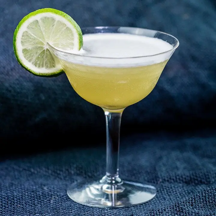 An elegant cocktail coupe rests on a blue canvas surface and backdrop. The drink within is bright yellow with a thin white foam head, garnished with a whole lime wheel.