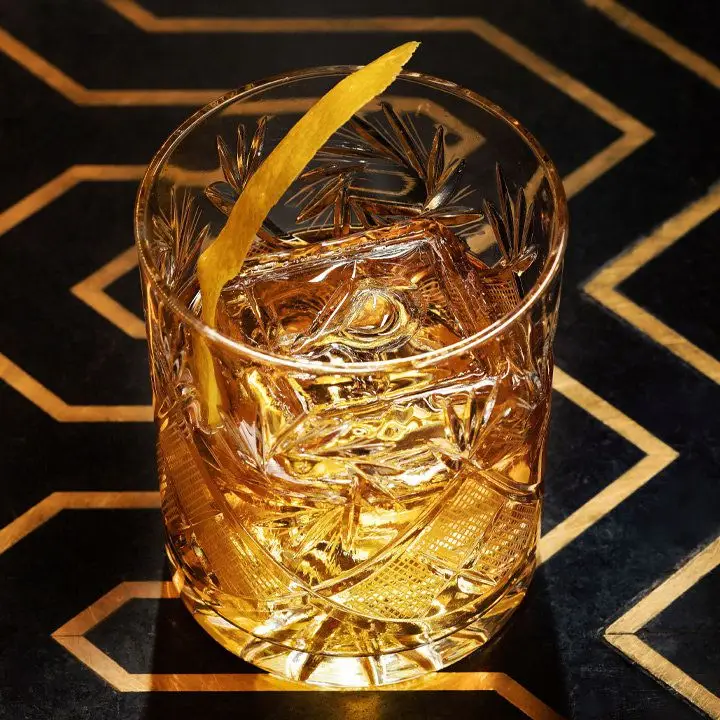 An intricately designed double Old Fashioned glass rests on a black surface with golden zig zagging inlays. The glass holds a large ice cube, a light brown drink, and a lemon zest.