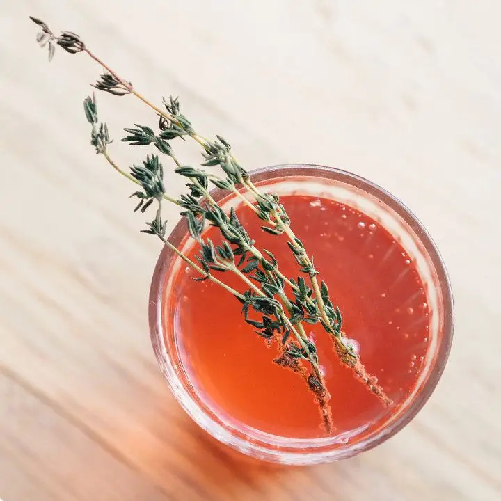 An overhead shot shows a highball glass resting on a pale hardwood surface. The glass is filled with a bubbly red-orange drink and garnished with two sprigs of thyme. 