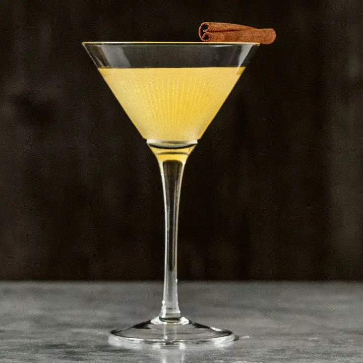 A delicate cocktail glass rests on a polished marble surface. It’s filled with a golden drink and garnished with a cinnamon stick.