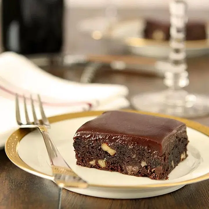 A brownie rests on a white plate with a gold rim. The brownie is filled with nuts and topped with a shiny glaze. An elegant fork rests next to it on the plate, and a napkin is hazy in the background.