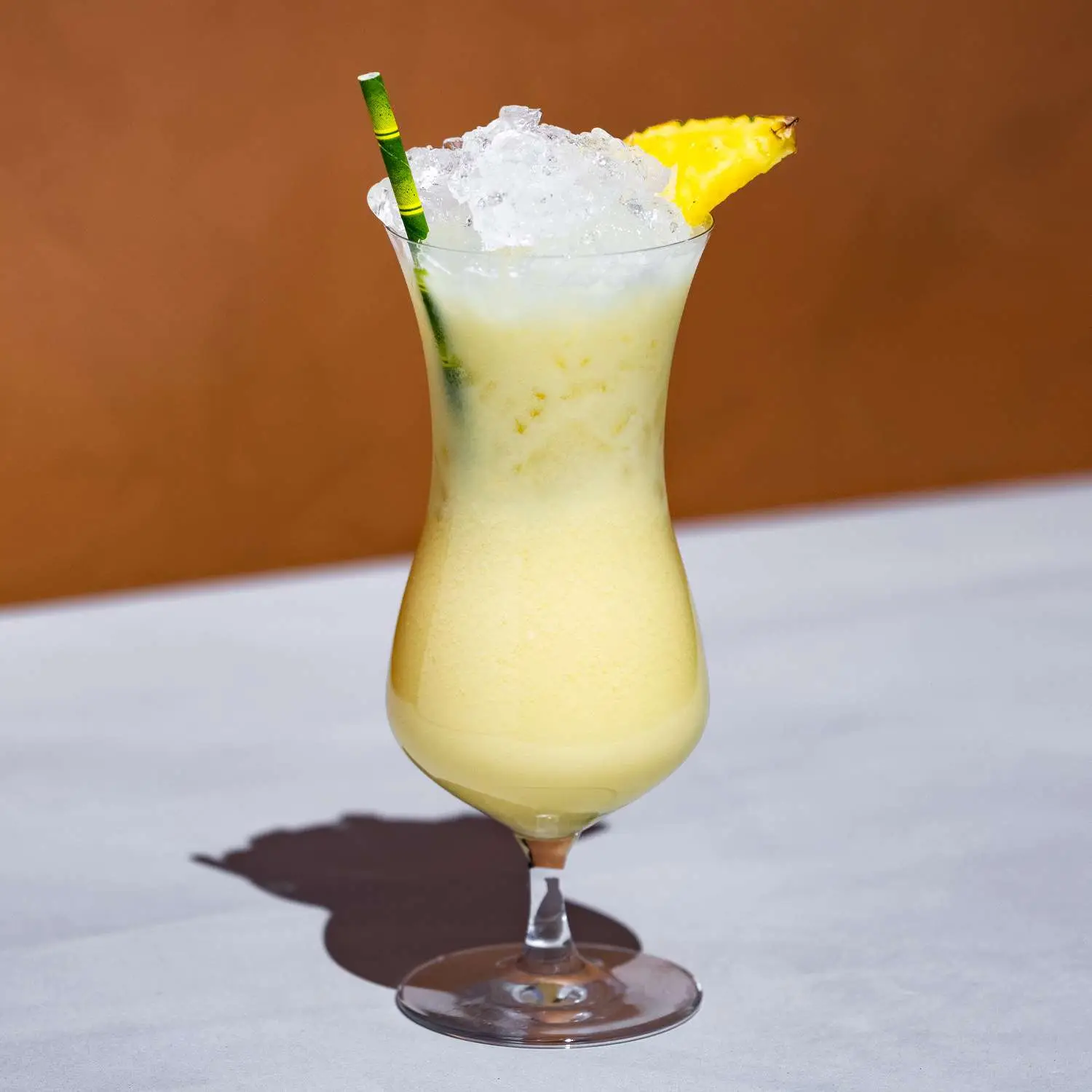Painkiller cocktail, pale yellow, in a curved hurricane glass over crushed ice, with a bamboo straw and pineapple wedge garnish