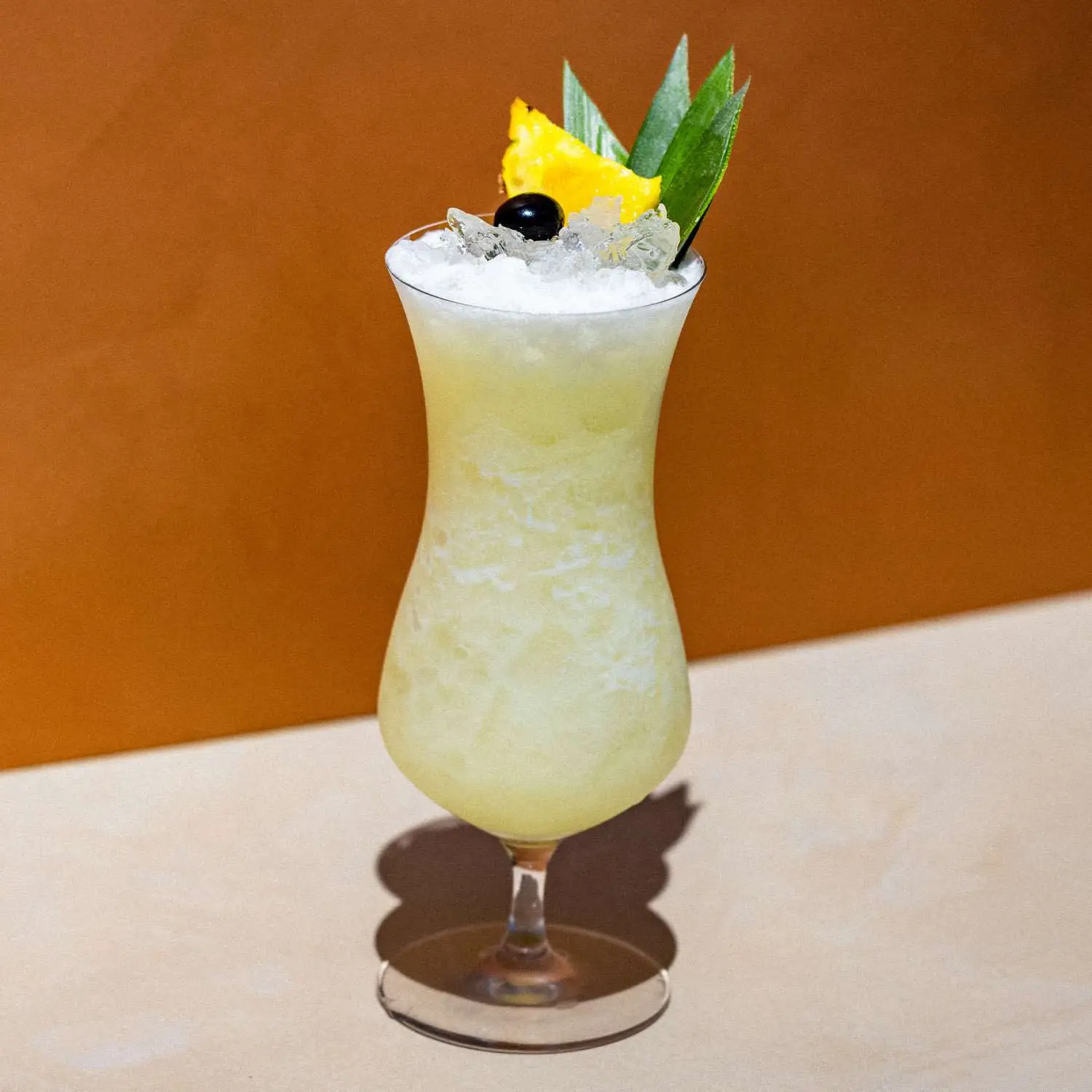 Frozen Pina Colada in curved hurricane glass with blended ice, garnished with pineapple wedge, leaves, and a maraschino cherry