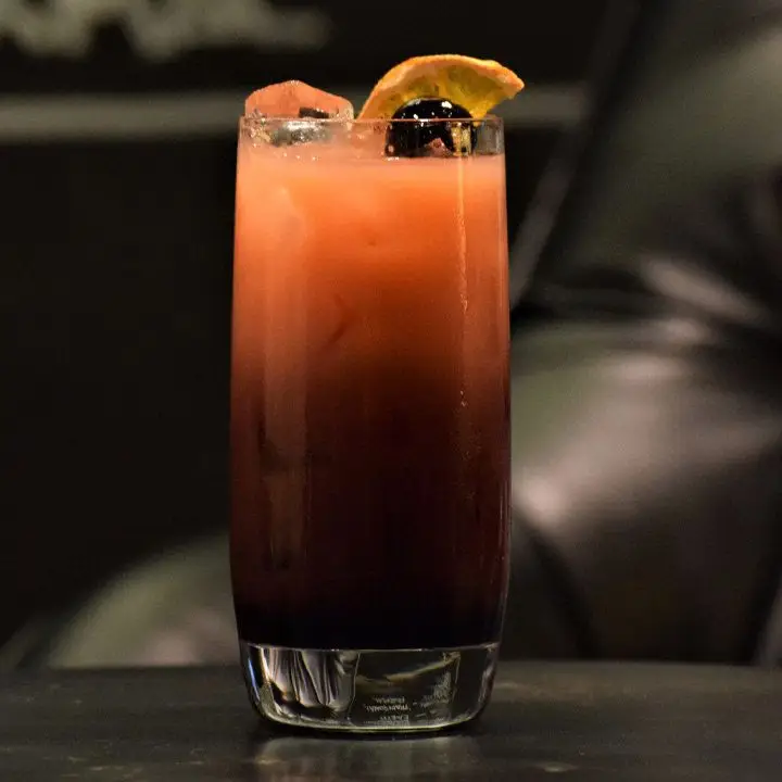 A Collins glass rests on a black surface in front of a black leather chair. The drink within is dark red at the base and lightens to orange as it gets to the top. 