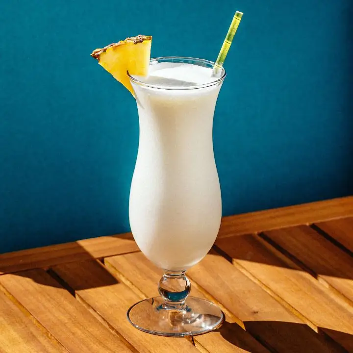 A curvy Hurricane glass rests on a slatted wooden surface against a sea blue wall. The glass holds a pale white blended coconut cocktail and is garnished with a pineapple wedge and a yellow and green straw. 