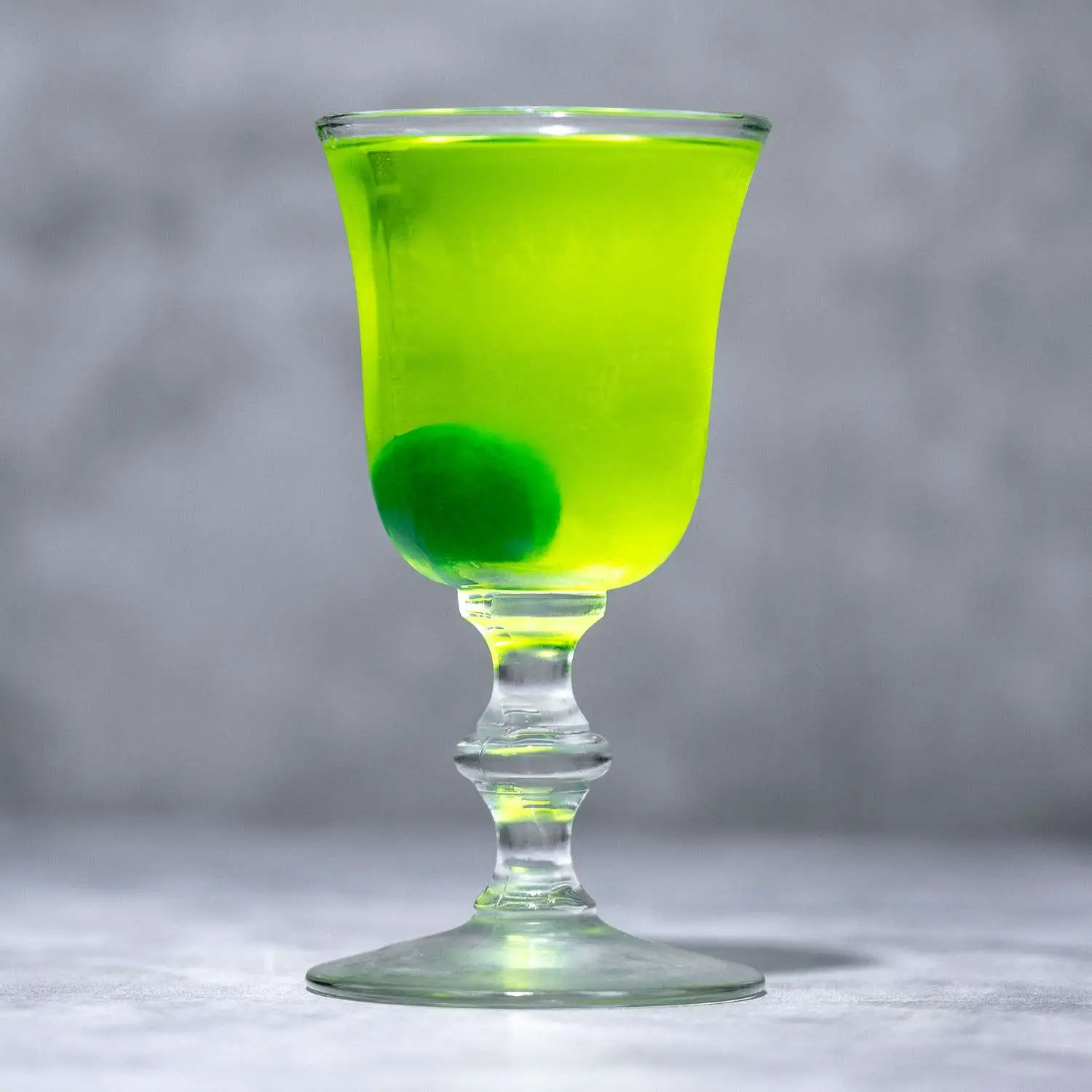 Neon green Greenhorn cocktail in Nick & Nora glass, with green maraschino cherry at bottom, on marble background