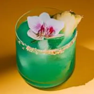A beautiful blue-green cocktail in a rocks glass is garnished with a lovely pink orchid and a slice of pineapple.