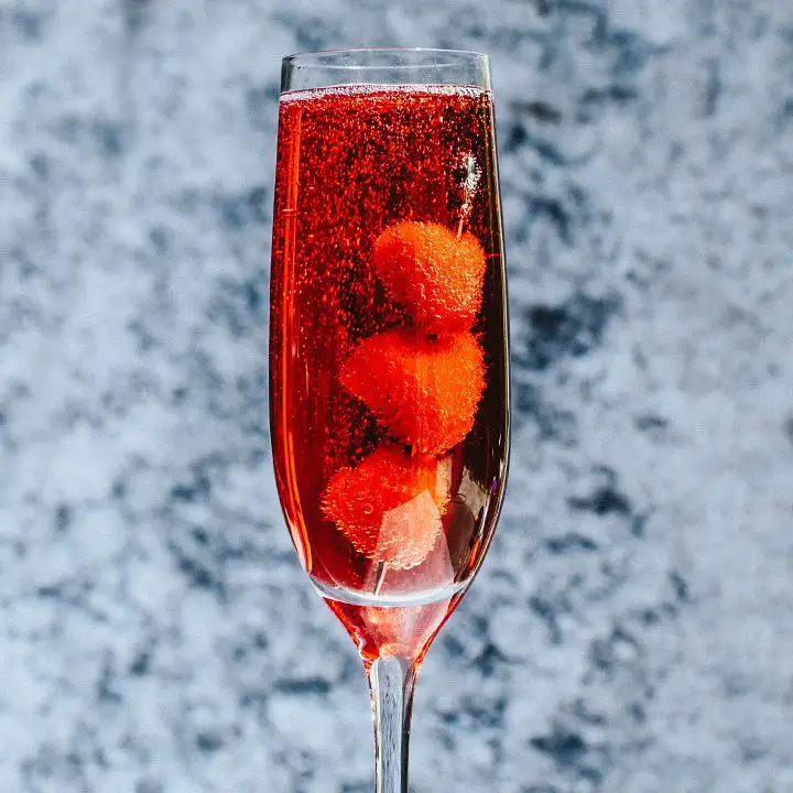 A stemmed Champagne flute bubbles with a crimson colored drink. Three berries on a silver skewer rest in the glass. The background is out of focus gray and blue mottled wallpaper.