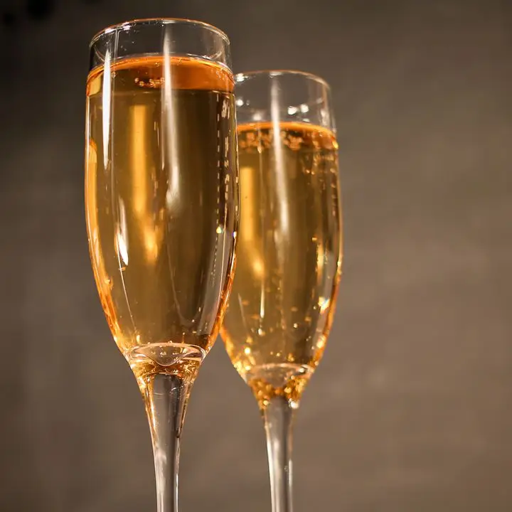 Two Champagne flutes are filled with a golden sparkling drink, set against a brown backdrop. 