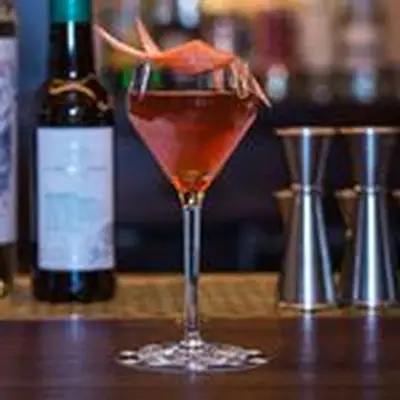 A cordial glass rests on a bar top with bottles and jiggers in the background. The drink within is red, and garnished with a curl of grapefruit peel.