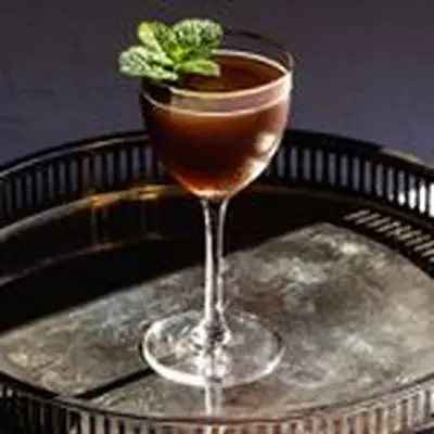 A Nick & Nora glass with a copper rim rests on an elegant bar tray. The glass holds a brown, opaque cocktail.