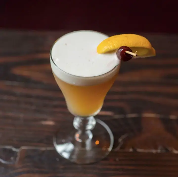 A sour glass rests on hardwood and holds a beautiful Whiskey Sour, complete with a thick layer of white foam, as well as a lemon wedge garnish and a cherry on a wooden skewer.