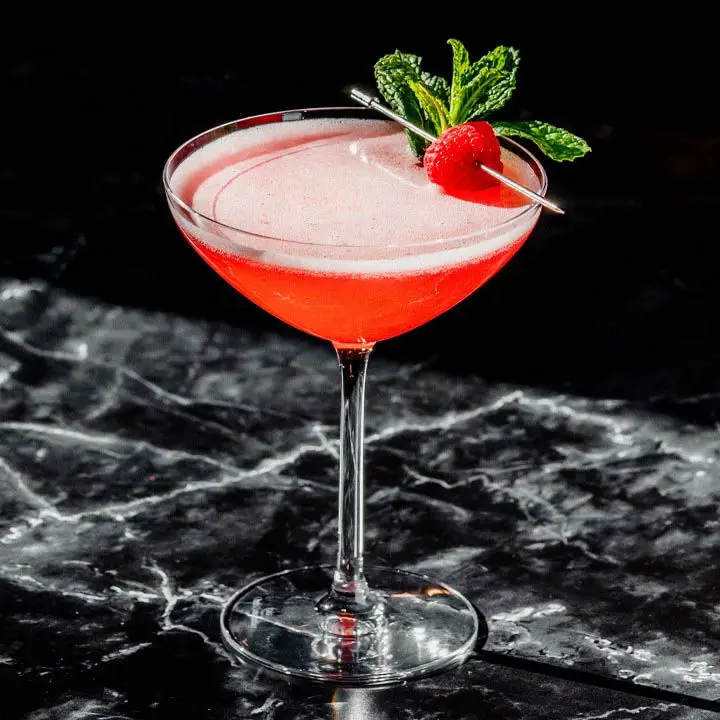 A wide, shallow and slightly curved Martini glass holds a vividly red cocktail. The drink is garnished with a sprig of mint and a raspberry on a silver pick. The surface below is black marble, the background solid black.