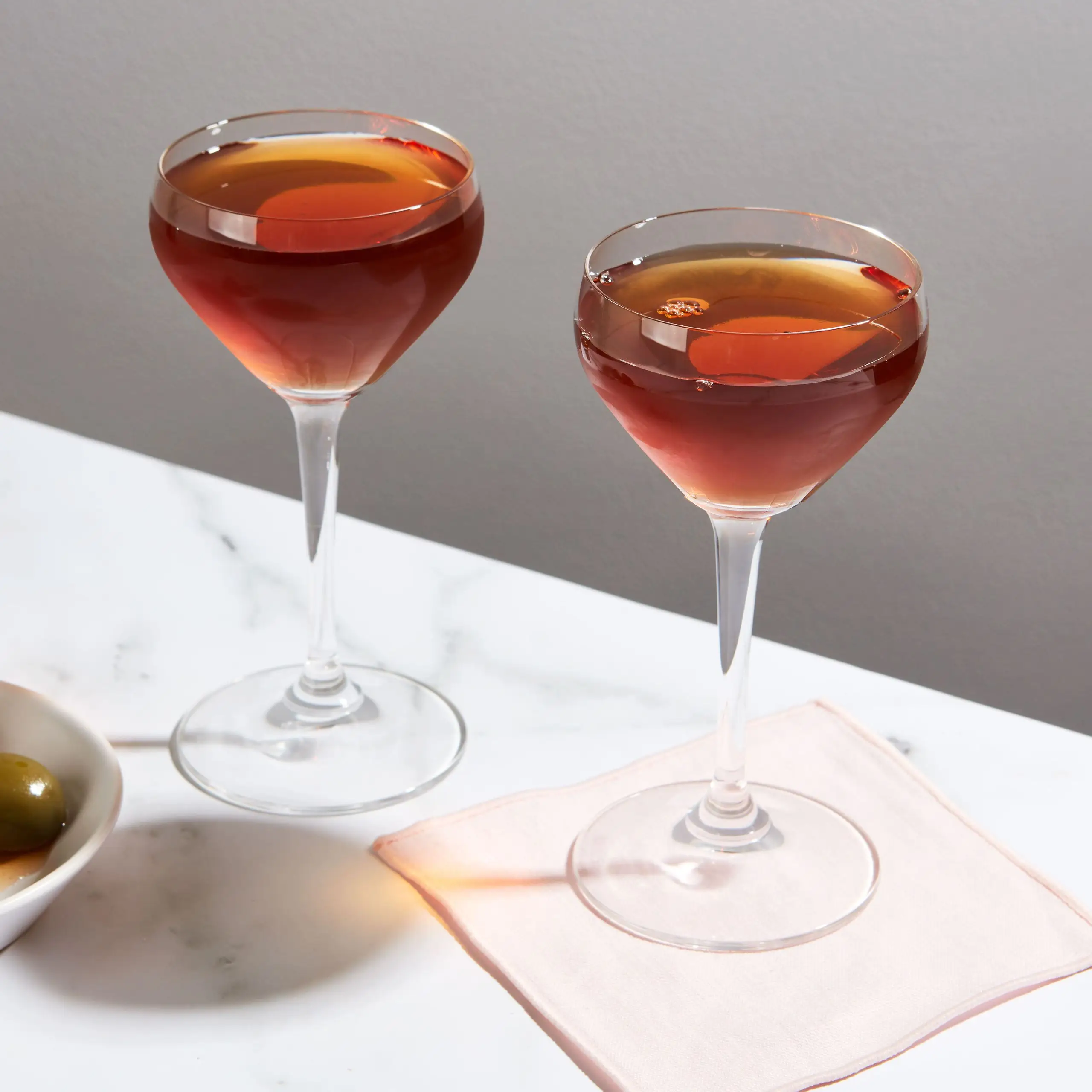 Photo of two glasses of an Adonis cocktail on a marble countertop with a cocktail napkin and a bowl of olives..