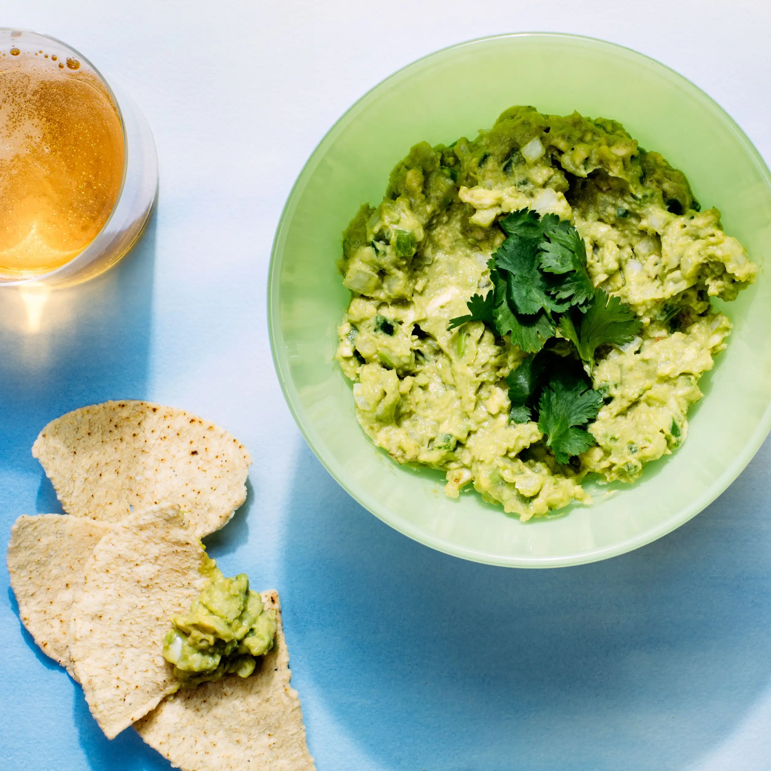 The best guacamole recipe in a green bowl topped with fresh cilantro. On the side are a bowl of tortilla chips and...