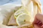 A salmon filet in a piece of parchment paper with snow peas underneath and slices of lemon on top.' title='Salmon en Papillote